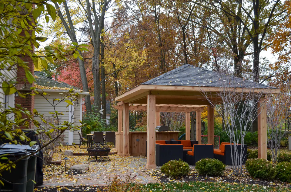 Freestanding covered pavilion in a fall backyard with lounge seating, paver patio, shrubs, and mature trees.
